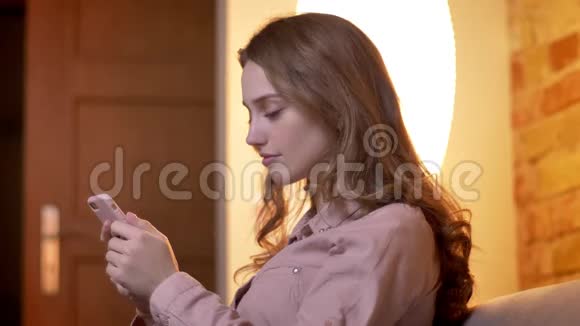 在舒适的家庭氛围中年轻少女观看智能手机的侧写变成相机和微笑视频的预览图