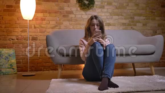 年轻女学生坐在地板上膝盖弯着腰在舒适的家着手机的画像视频的预览图