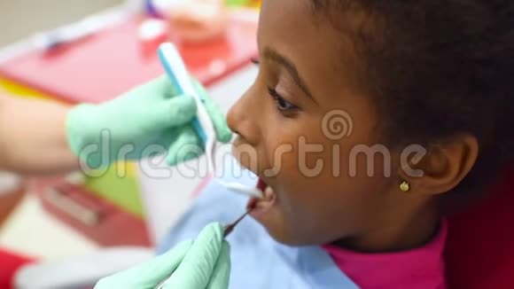 可爱的深色皮肤的非裔美国女孩坐在一个红色的黄色儿童牙科椅上看牙医视频的预览图