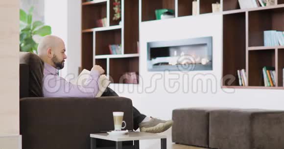 男人正在酒店大厅的咖啡馆里读一本书卡布奇诺杯坐在壁炉前的沙发上视频的预览图