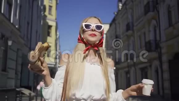 积极美丽的年轻女子走在街上喝咖啡吃牛角面包漂亮的时髦女孩视频的预览图