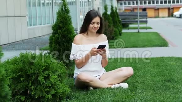 年轻漂亮的女人坐在酒店附近的草地上使用智能手机视频的预览图
