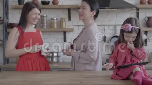 祖母母亲和小女儿在一个现代化的厨房里那个拿着平板电脑坐在桌子上的女孩视频的预览图