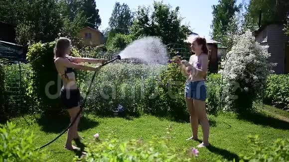 夏天阳光明媚的一天两个年轻漂亮的女孩从花园的软管里互相倒在绿色的草坪上穿短短裤的金发美女视频的预览图
