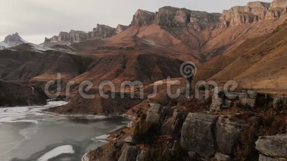一个戴着兜帽的人站在靠近山湖的悬崖边上以史诗般的岩石为背景视频的预览图