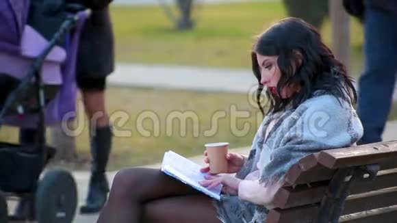 漂亮可爱的黑发女孩坐在城市公园的长凳上喝热咖啡里面有蒸汽和读书视频的预览图