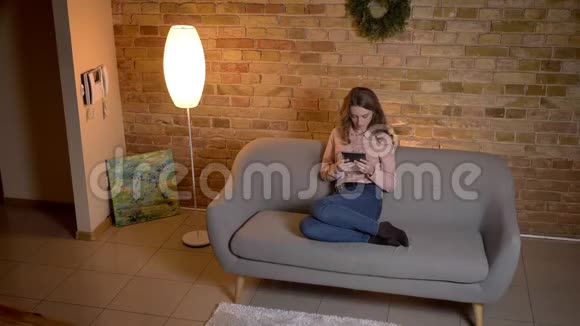 在舒适的家中坐在沙发上和平板电脑一起工作的高浓缩金发少女的顶级镜头视频的预览图