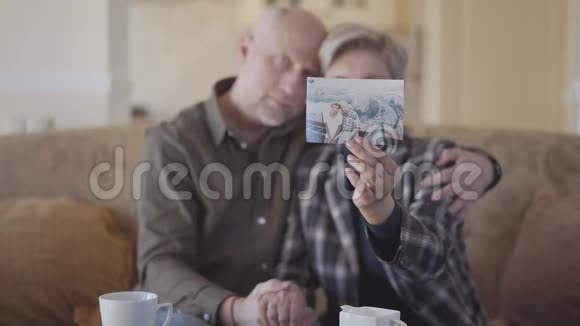 成年夫妇坐在沙发上展示他们的旧照片分享他们的记忆焦点从照片转移到已婚视频的预览图