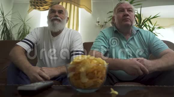 两个英俊的高级男人坐在棕色的皮革沙发上看电视退休人员的休闲视频的预览图