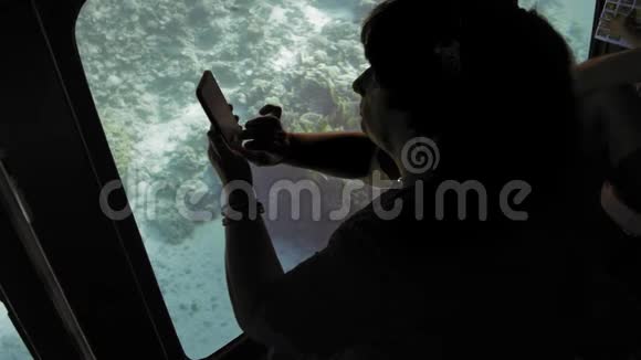 在一艘潜水艇上观看海底珊瑚礁的资深成年女性游客在智能手机上拍摄鱼视频的预览图