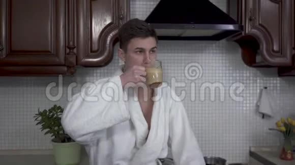 自信的男人穿着白色浴袍在现代厨房里喝咖啡卡布奇诺年轻人把透明的杯子放在桌子上视频的预览图