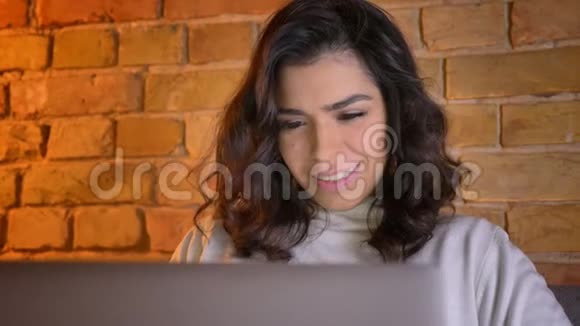 一幅充满欢乐的白种人黑发女商人的画像她坐在沙发上微笑着笔记本电脑视频的预览图