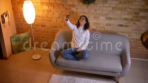 漂亮的黑发家庭主妇坐在沙发上在家里愉快地使用智能手机制作自拍照片视频的预览图