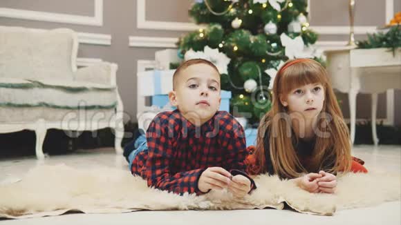 美丽的兄弟姐妹在圣诞杉木下躺在地毯上看新年电影的温暖照片视频的预览图