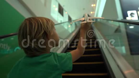 一个小男孩手里拿着白色玩具飞机在自动扶梯上移动的慢镜头自由概念视频的预览图