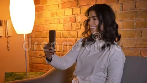 一位白种人黑发家庭主妇坐在沙发上在智能手机上愉快地用视频聊天她的特写肖像视频的预览图