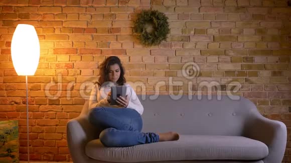 一幅漂亮的黑发家庭主妇坐在沙发上在家里兴高采烈地看着平板电脑的肖像视频的预览图