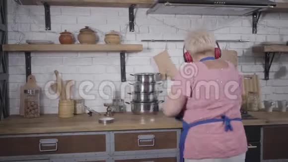 精力充沛的舞蹈成熟的白种人女人戴着耳机在厨房做饭有趣的高级夫人把意大利面视频的预览图