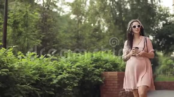 怀着幸福的年轻女人手里拿着咖啡在城市公园散步视频的预览图