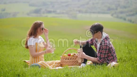 一对年轻夫妇穿着鲜艳的衣服坐在一片明亮的绿色草地上酒杯里倒着酒视频的预览图