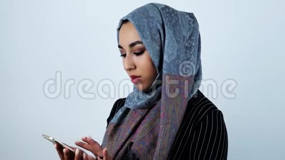 年轻漂亮的阿拉伯女人一边微笑一边在白色背景上滚动她的智能手机视频的预览图