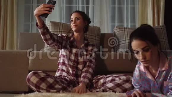 两个漂亮的姐妹之一在智能手机上自拍而另一个则在晚上舒适的客厅里阅读杂志视频的预览图