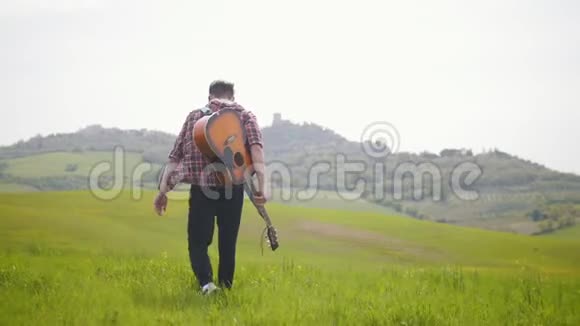 一位年轻的吉他手走在明亮的绿色草地上视频的预览图