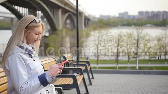 年轻漂亮的金发女郎在智能手机上写信息一个年轻的女人正在河堤的长凳上休息视频的预览图