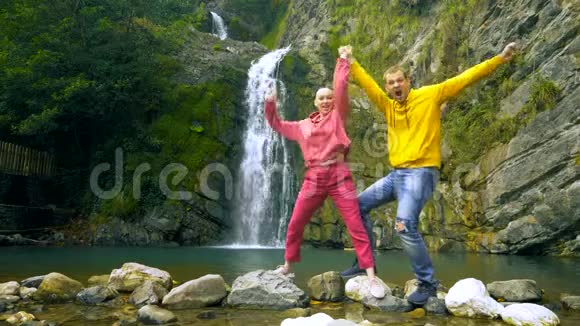 一对夫妇一个年轻人和一个秃头女孩正在瀑布的背景下举起手臂跳舞视频的预览图