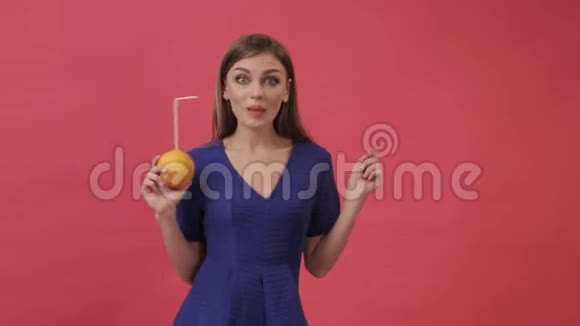 漂亮的女孩穿着裙子喝橙汁用鸡尾酒吸管跳舞紫色背景工作室视频的预览图
