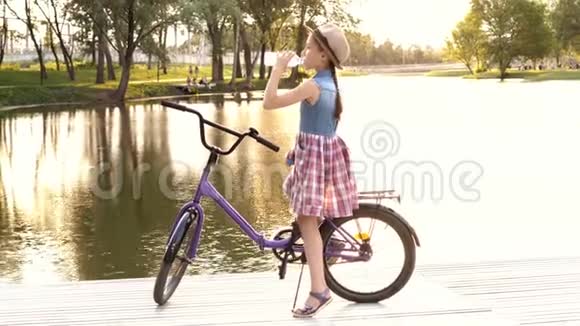 日落时分一个小女孩坐在河边的一辆紫丁香自行车上从一个透明的瓶子里喝水明亮明亮视频的预览图