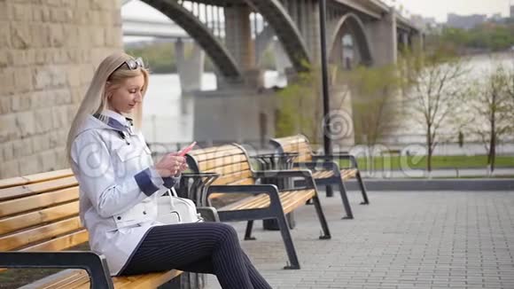 年轻漂亮的金发女郎在智能手机上写信息一个年轻的女人正在河堤的长凳上休息视频的预览图
