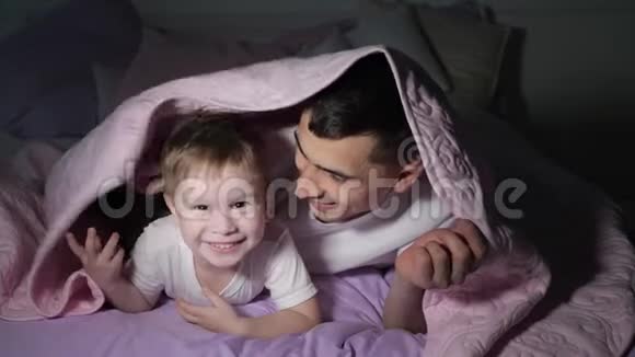 爸爸和他的小儿子在黑暗中躲在毯子下面玩视频的预览图