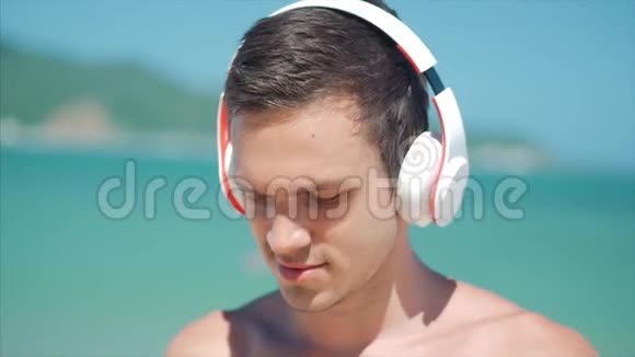 拥抱英俊的年轻人用无线白耳机听他的智能手机音乐在海滩上跳舞视频的预览图