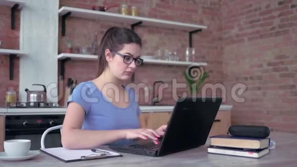 年轻的现代女性在家工作戴眼镜的女孩在手提电脑键盘上打字坐在剪贴板上写笔记视频的预览图