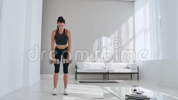 一位年轻的运动女子在一间明亮的公寓里用哑铃做一次举重使她向前倾斜视频的预览图