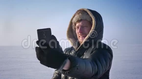 穿着保暖外套和引擎盖的商人在雪地沙漠的智能手机上有一个视频卡的特写肖像视频的预览图