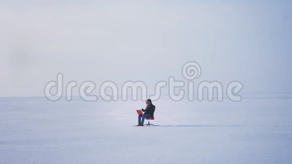 一个穿着温暖外套和引擎盖的商人坐在雪地沙漠里专心地用笔记本电脑工作视频的预览图