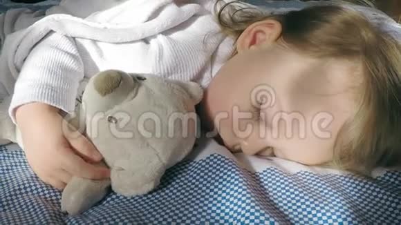 美丽的小女孩金发睡在床上手里拿着一只玩具熊被太阳照亮视频的预览图