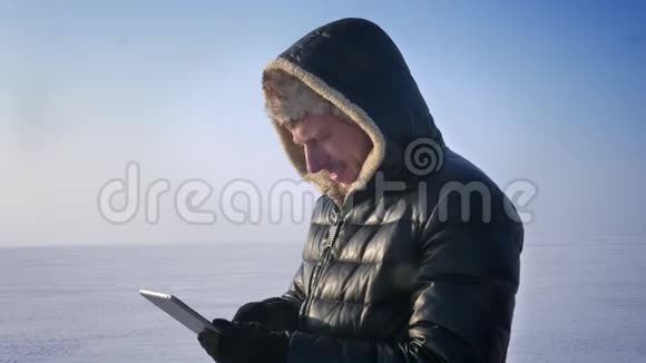 商人穿着温暖的外套和引擎盖站在冰冻的湖面上用平板电脑工作认真地转向相机和手表视频的预览图