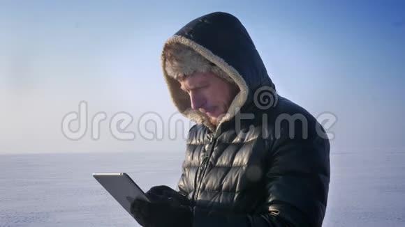 商人穿着温暖的外套和引擎盖站在冰冻的湖面上用平板电脑工作转向相机兴高采烈地微笑视频的预览图