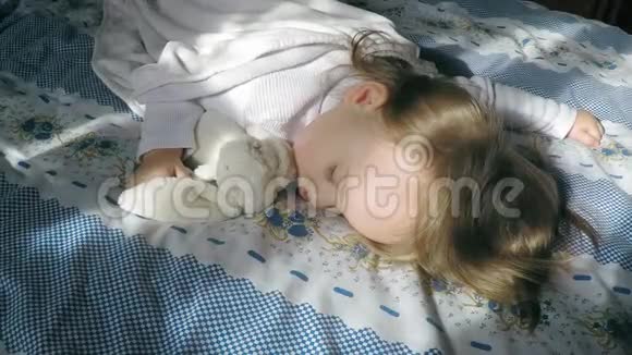美丽的小女孩金发睡在床上被阳光照亮抱着一只毛绒兔子视频的预览图