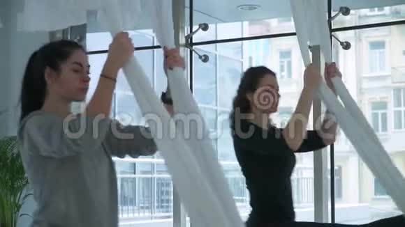 两个女人在吊床上徘徊在室内工作室放松瑜伽运动女孩练习空中瑜伽视频的预览图