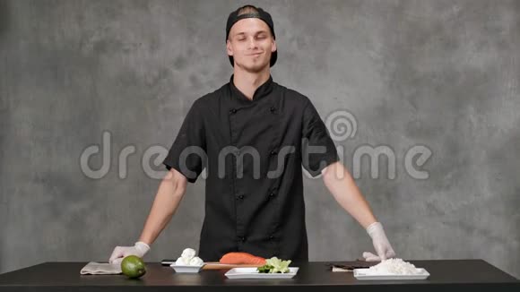 年轻的厨师穿着黑色衣服在灰色背景下做饭厨房餐桌日本寿司和卷的原料视频的预览图