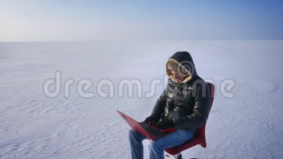 穿着温暖外套和引擎盖的商人坐在雪地里拿着笔记本电脑兴高采烈地伸展着自己的肖像视频的预览图
