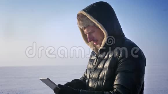 商人穿着温暖的外套和引擎盖站在冰冻的湖面上展示着绿色的平板电脑屏幕推荐这款应用程序视频的预览图