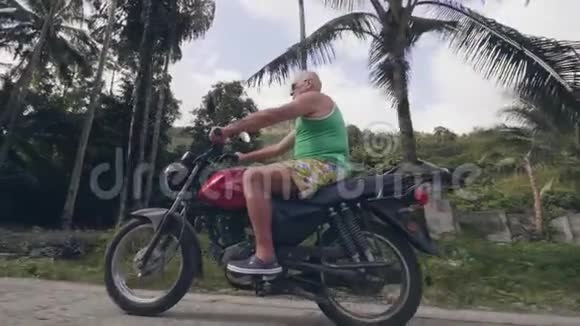 成熟的人骑摩托车在村道上的热带棕榈树景观骑摩托车的老人视频的预览图