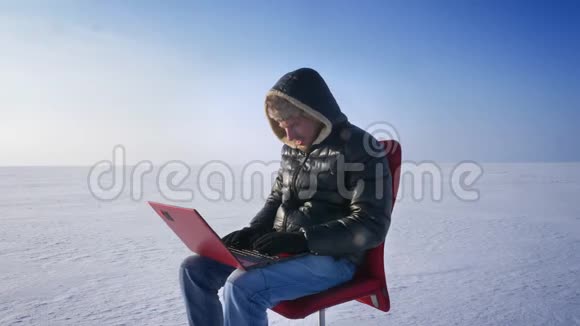 孤独的商人穿着温暖的外套和引擎盖坐在雪地沙漠里用心地用笔记本电脑工作的肖像视频的预览图