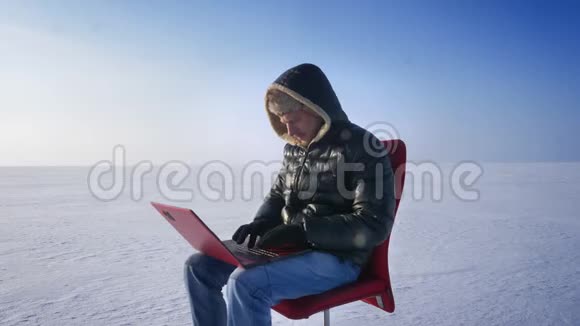穿着温暖外套和引擎盖的商人坐在雪地沙漠里用笔记本电脑工作转向相机兴高采烈地微笑着视频的预览图