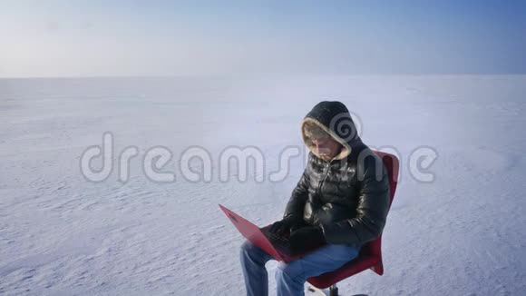 孤独的商人穿着温暖的外套和引擎盖坐在雪地沙漠里用笔记本电脑和伟大的笔记本电脑工作的肖像视频的预览图